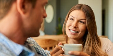A woman and a man sitting next to each other. The woman has a cup in her hands and smiles at the guy.