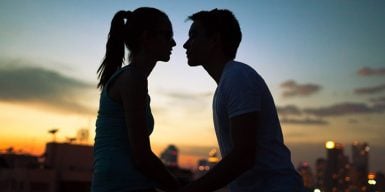 A couple sitting at the sunset, leaning in on each other for a kiss.