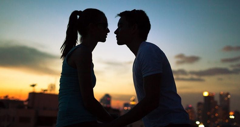A couple sitting at the sunset, leaning in on each other for a kiss.