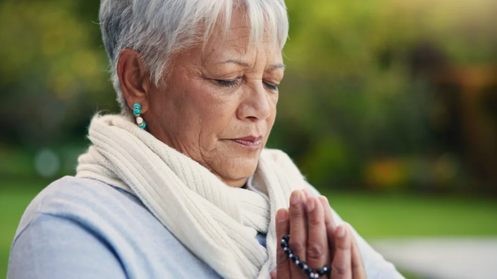 Elderly woman praying with rosary beads outdoors.