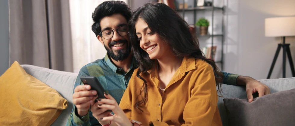 A couple sitting on their couch together an looking at a phone together.