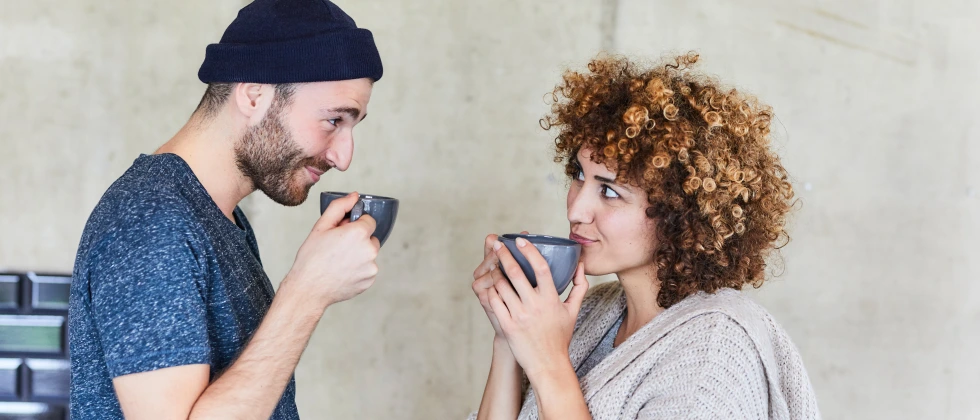 A guy and a woman drinking coffee, looking at each other in a flirty way, showing is it okay to ask out a coworker.