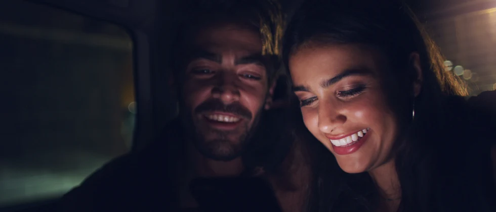 A couple in the back of a car smiling and looking at a phone.