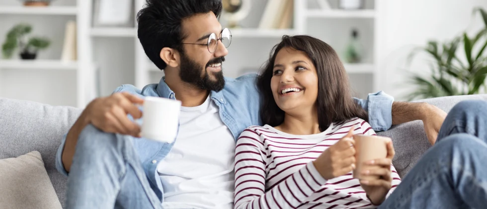 A couple on a couch holding cups and talking.